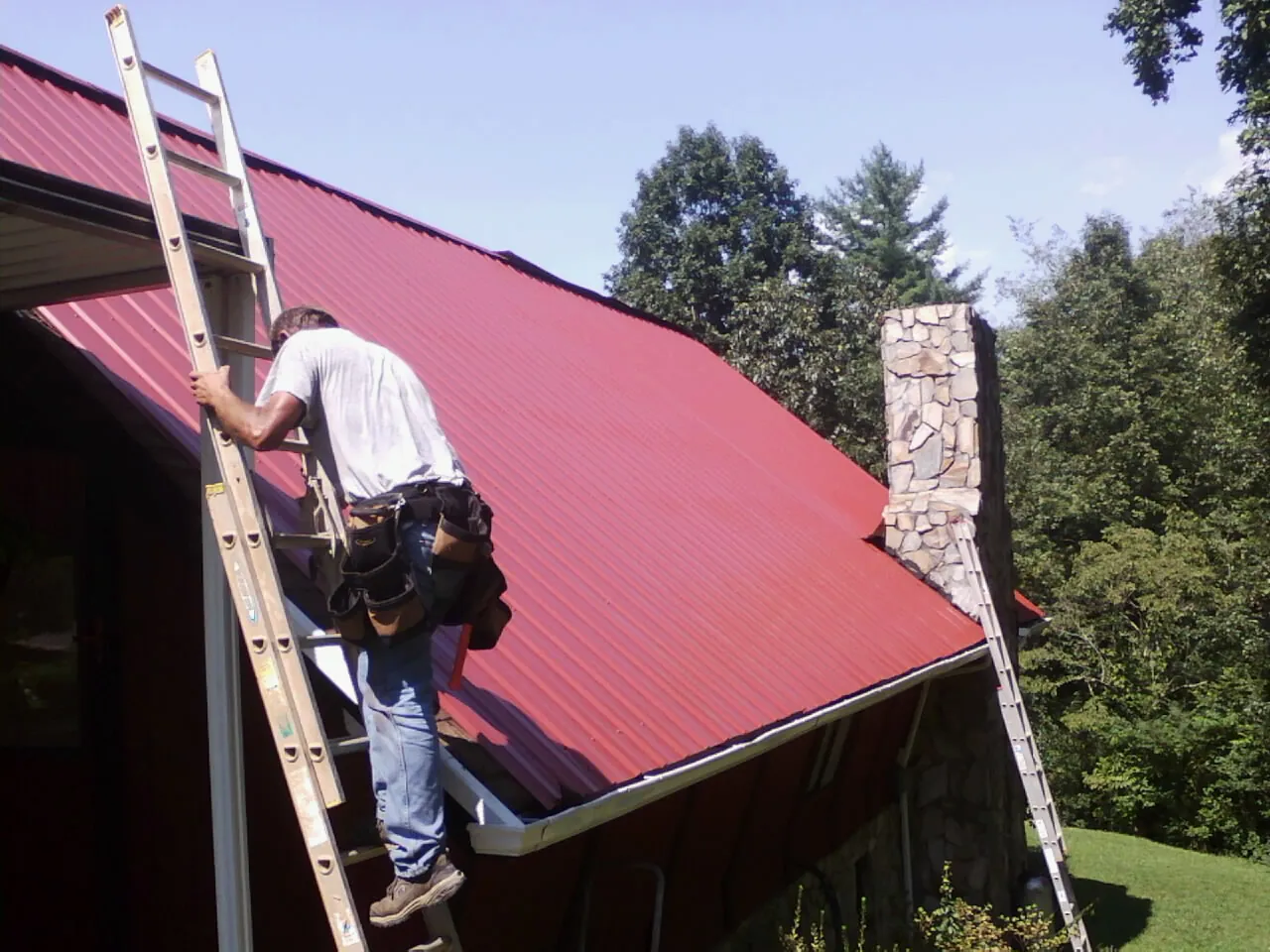 Roofer on a ladder beside a red metal roof and stone chimney — Soffit & Fascia Repair by Roofing Brothers in Mulvane
